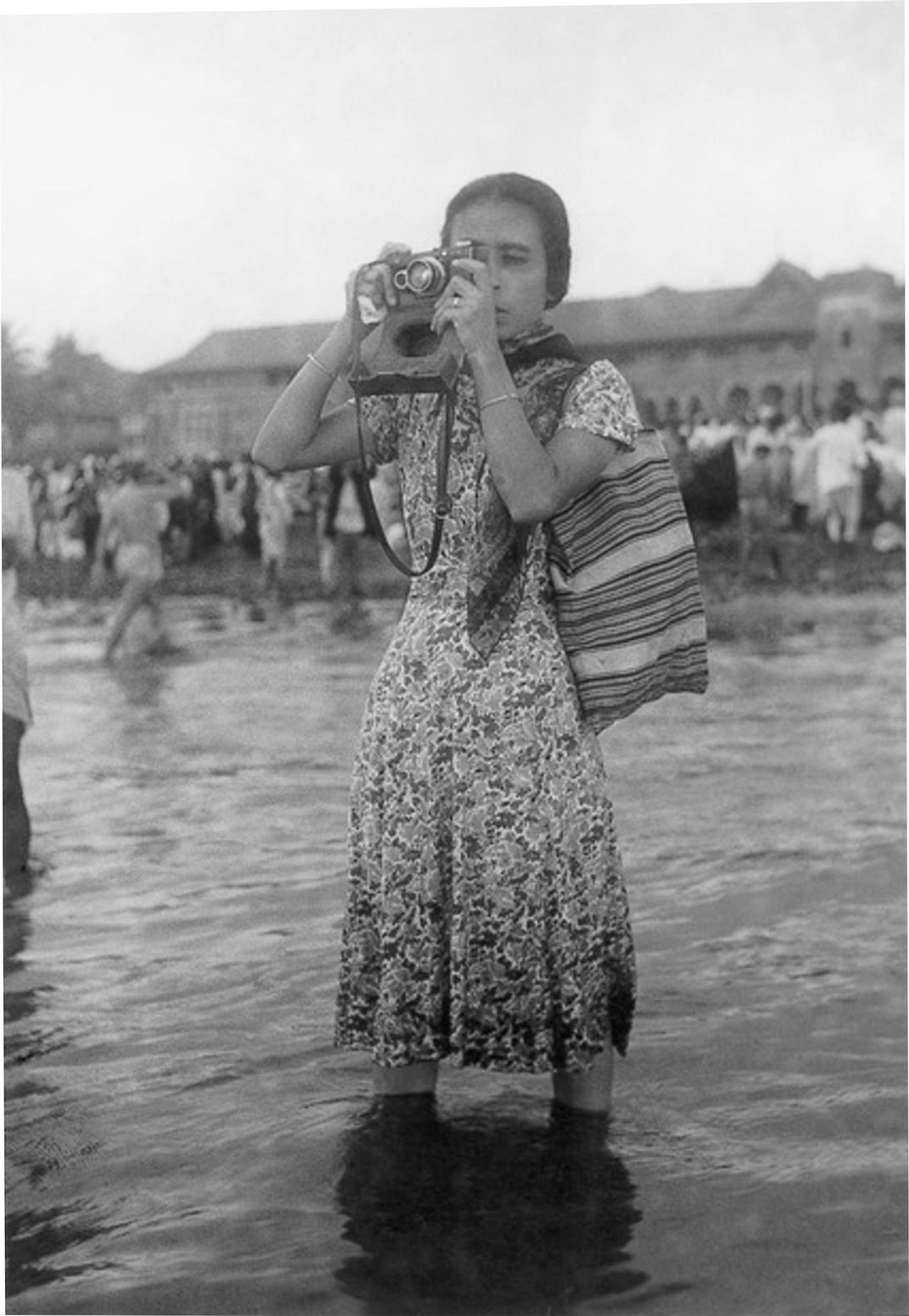 A black and white image of a woman in a dress standing ankle-deep in water, photographing something out of view of the camera