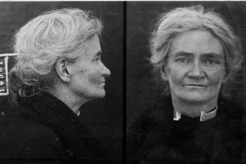 A black and white criminal mugshot of a white woman with graying hair, in a black outfit