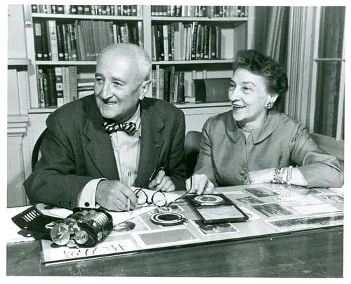 William, left, a white balding man in a bowtie and suit, and Elizebeth, right, a white woman in a suit jacket, sit at a desk with codebreaking materials in front of them; both are elderly