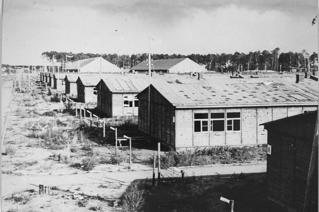A black and white image of low rectangular buildings with peaked roofs, grassy dirt ground, black windows and shadows