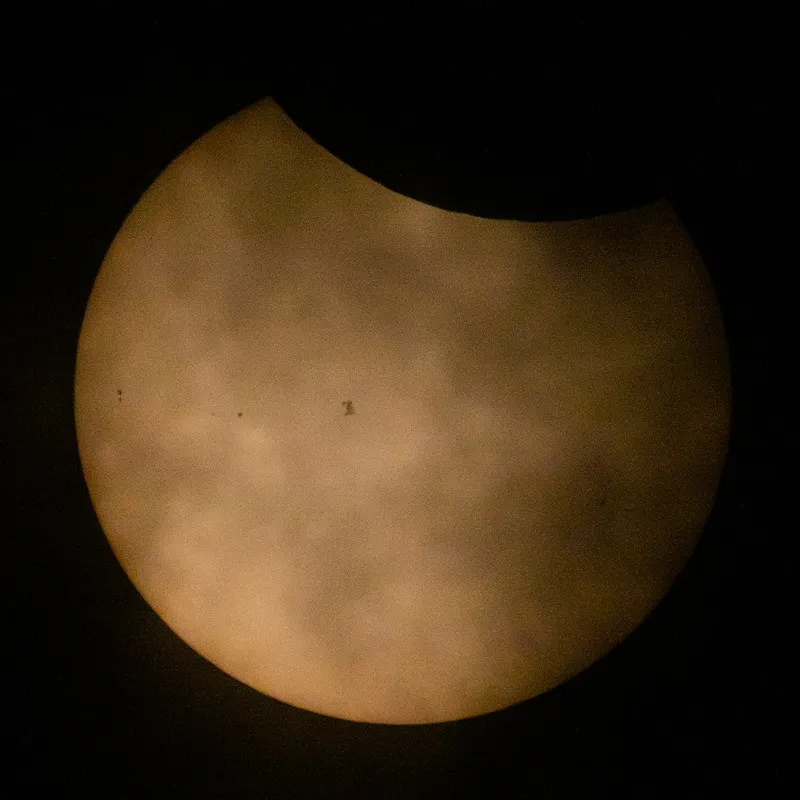 the surface of the sun looks like a dim orange orb, with the moon obscuring about ten percent at the top, and the silhouette of a spacecraft covers part of its surface