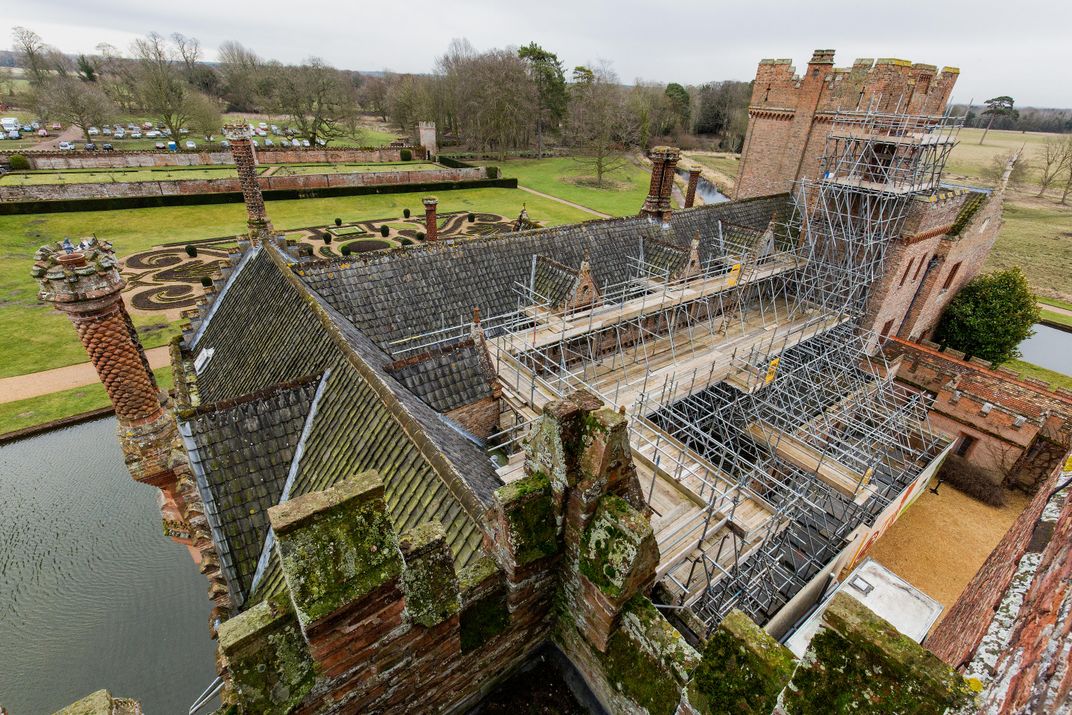 A view looking down on the roof of the Manor, with about half of its roof covered by scaffolding. Behind the house, which is covered in moss, there's a bit of moat and an ornate garden