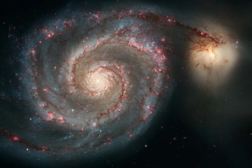 a swirling spiral galaxy with a yellow center and arms with blue and pink, one arm stretches and interacts with another galaxy