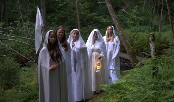 Five adults dressed in white dresses, robes, and veils pose along a forest path. One carries a lit lantern, and another carries a white flag.