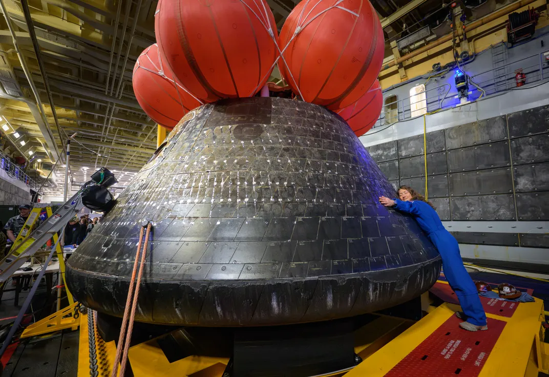 the Orion spacecraft indoors with its red balloons on top, and astronaut Christina Koch, wearing all blue, leans forward and gives it a hug
