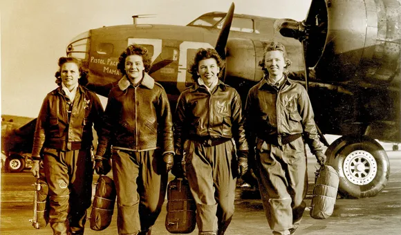 A black-and-white photo of four white women in their late 20s to early 30s walking on a military airfield. The women are wearing flightsuits, boots, and leather flight jackets. Behind the women is the forward fuselage of a World War II military bomber.