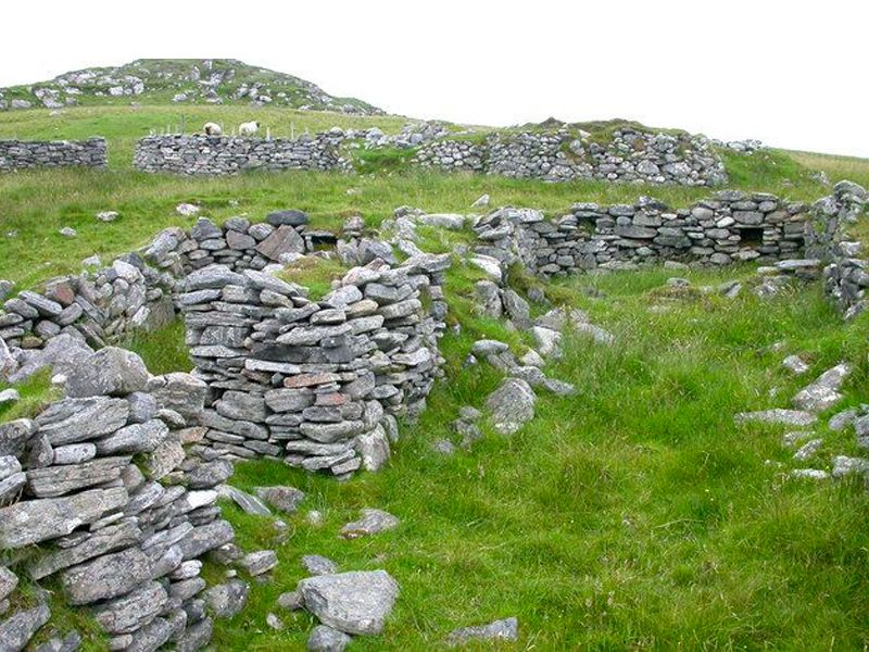 A field of green grass with a glimpse of grey sky behind; in the foreground, low stacked walls of flat rocks form the remains of terraces built into a sloping hill