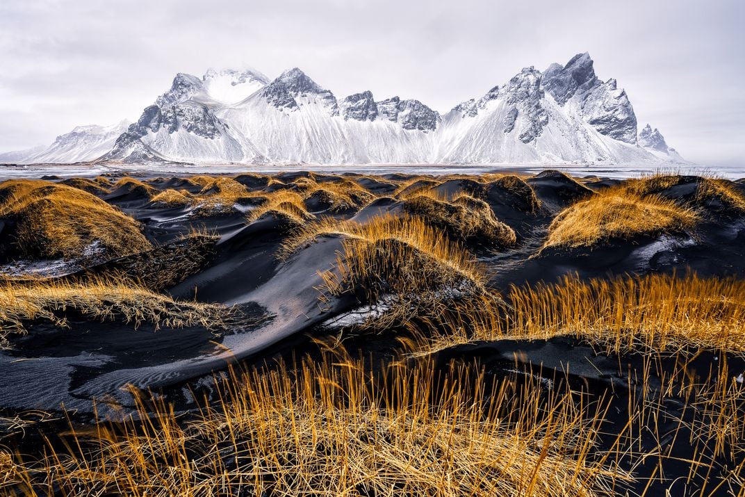 black sand and vegetation in front of icy mountains