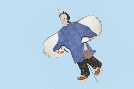 This paper kite depicts a Chinese girl wearing a long-sleeve blue tunic and loose-fitting black plants. A bamboo frame runs the length of the kite behind the paper.