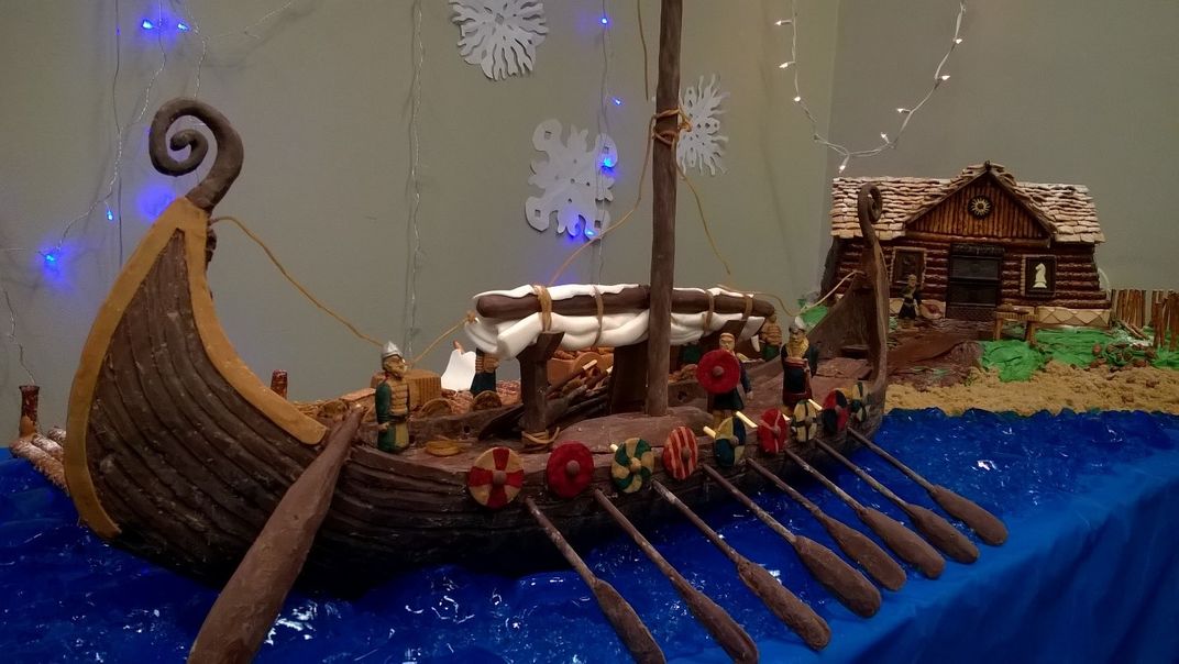 Brown, chocolate recreation of a viking ship complete with oars docked near a brown, chocolate cake recreation of a viking home on display on a rectangular table with a blue tablecloth.