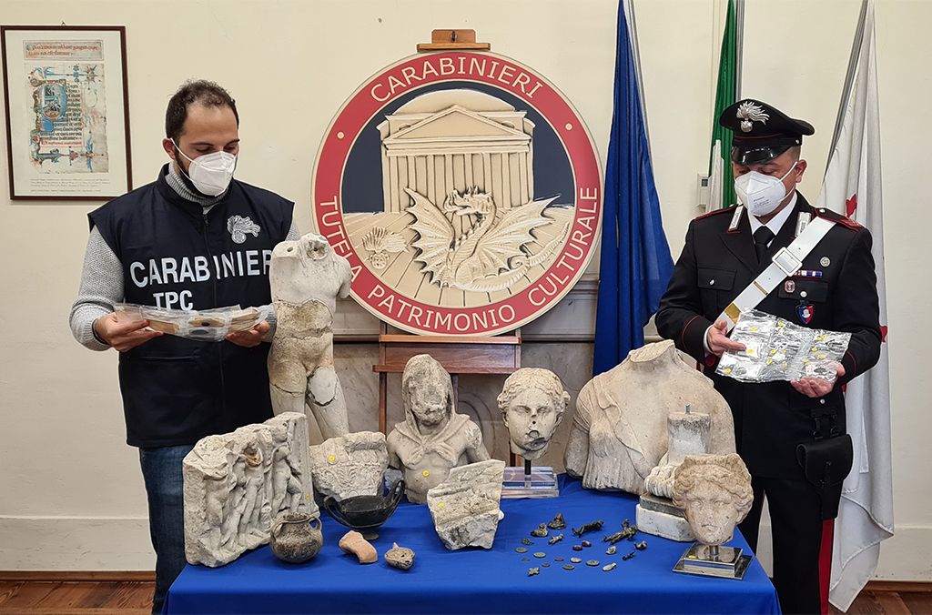 Two people in police uniforms stand behind a table covered in blue tablecloth, upon which sit several fragments of sculptures