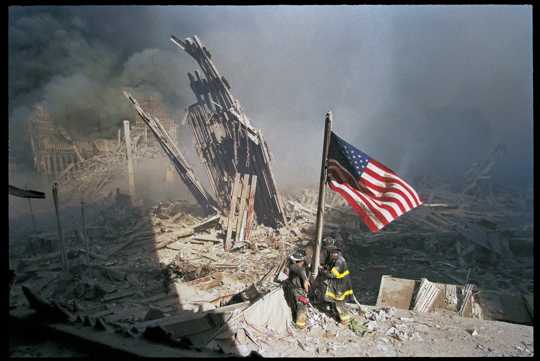 A dramatic horizontal view of the scene, with the shadow of a building cutting across the left side of the frame and the bright red stripes of the flag unfurling on its right, and an overwhelming amount of wreckage, dust and smoke in between