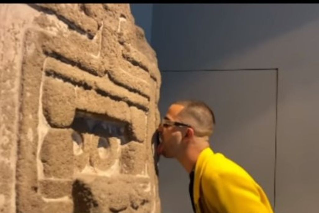 In a screenshot, a man in a yellow jacket licks the carved stone facade of an ancient work of Indigenous art