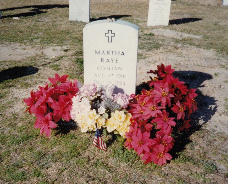 Martha Raye’s headstone decorated with flowers and a small U.S. flag. Other headstones in the Fort Bragg Main Post Cemetery are visible in the background.