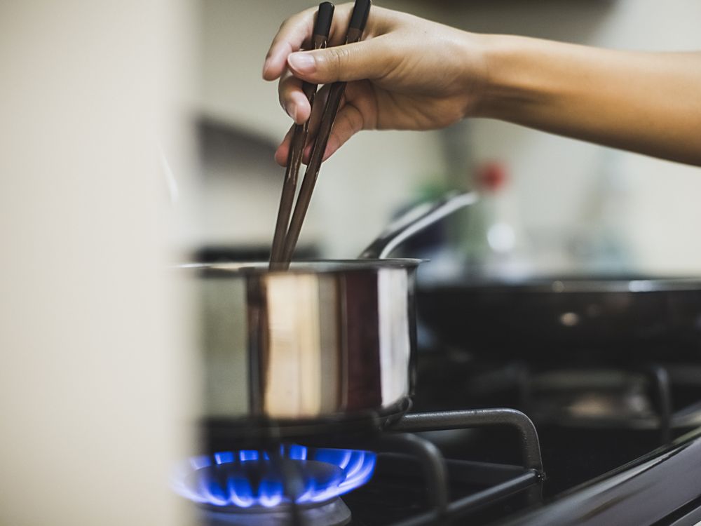 A woman's hand stirring a pot with chopsticks over a lit gas stove