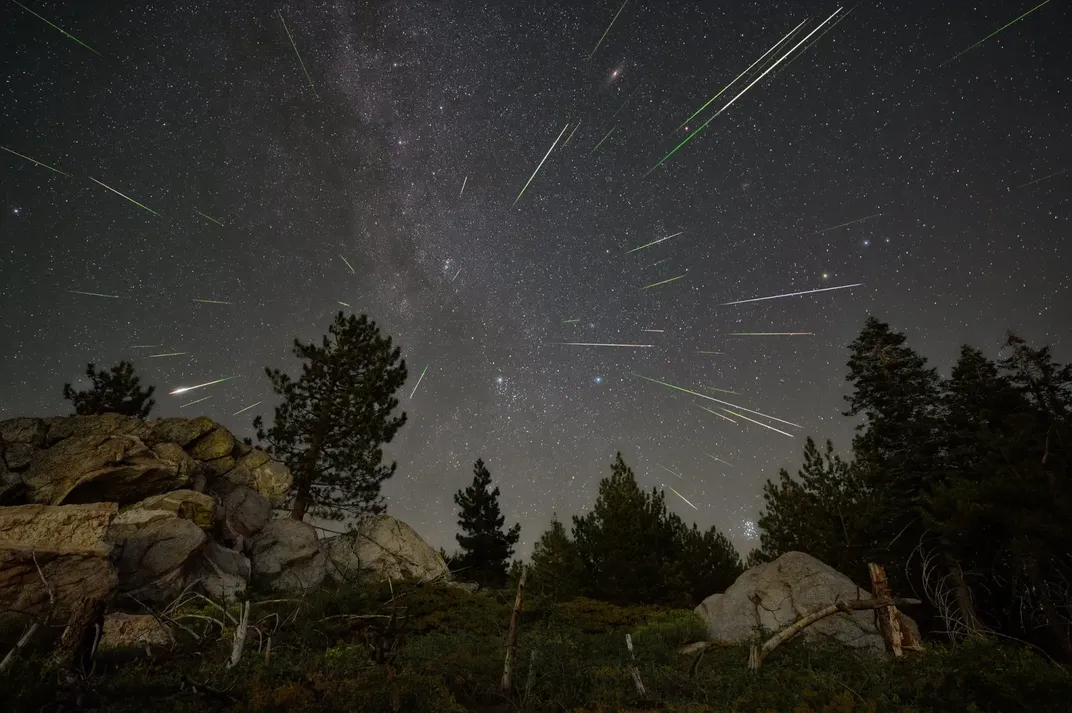 a couple dozen streaks of light emanate from the center of the image against a starry sky, with rocky cliffs and trees below