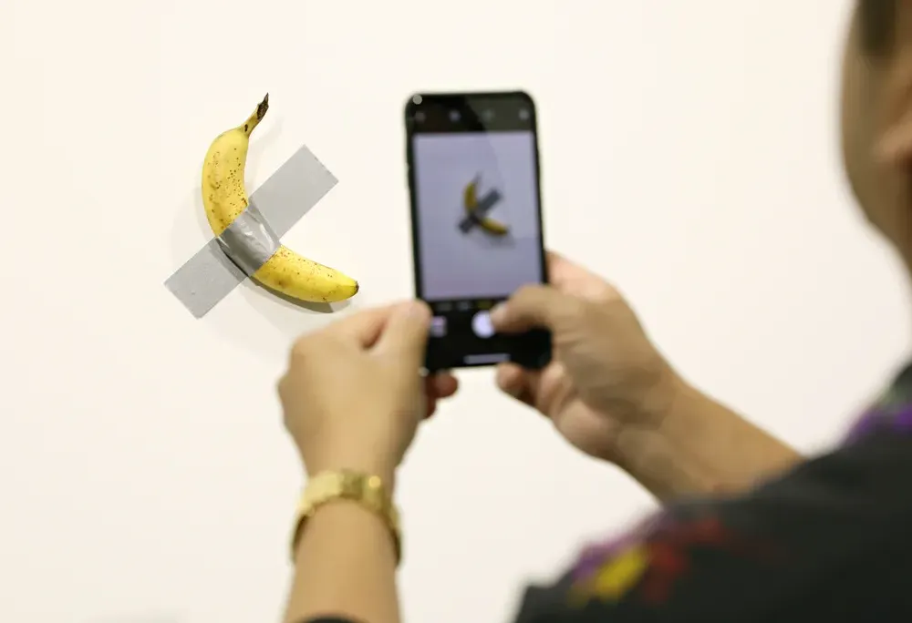 Person taking a photo of a banana taped to a white wall