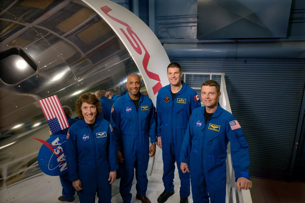 Four astronauts stand smiling in blue suits
