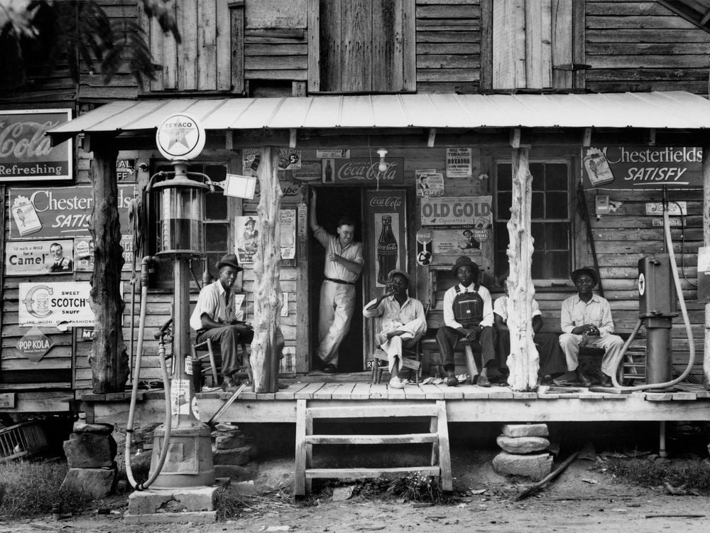 A group of people sit on the porch of a general store, surrounded by signs for products like Coca Cola and Chesterfield's cigarretes; most look at the camera 
