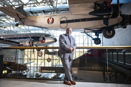 A Black man in his early 70s, wearing a blue suit and brown dress shoes, stands on a mezzanine overlooking a museum gallery displaying at least five airplanes.