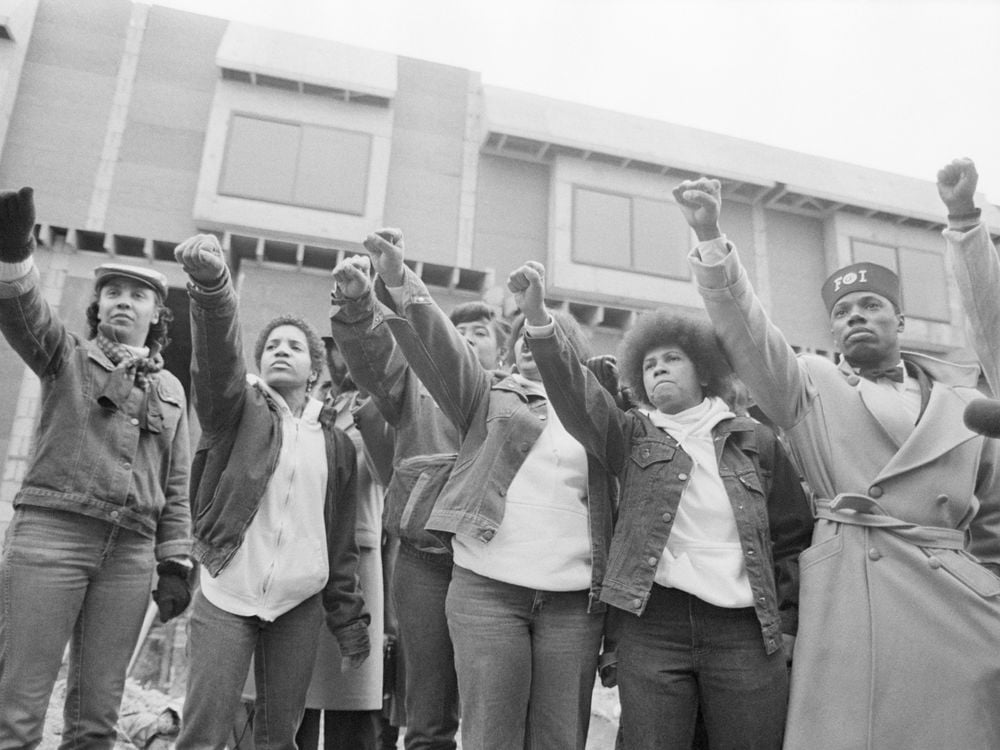 A black and white image of seven people, all Black, raising their right fists in the air in a line
