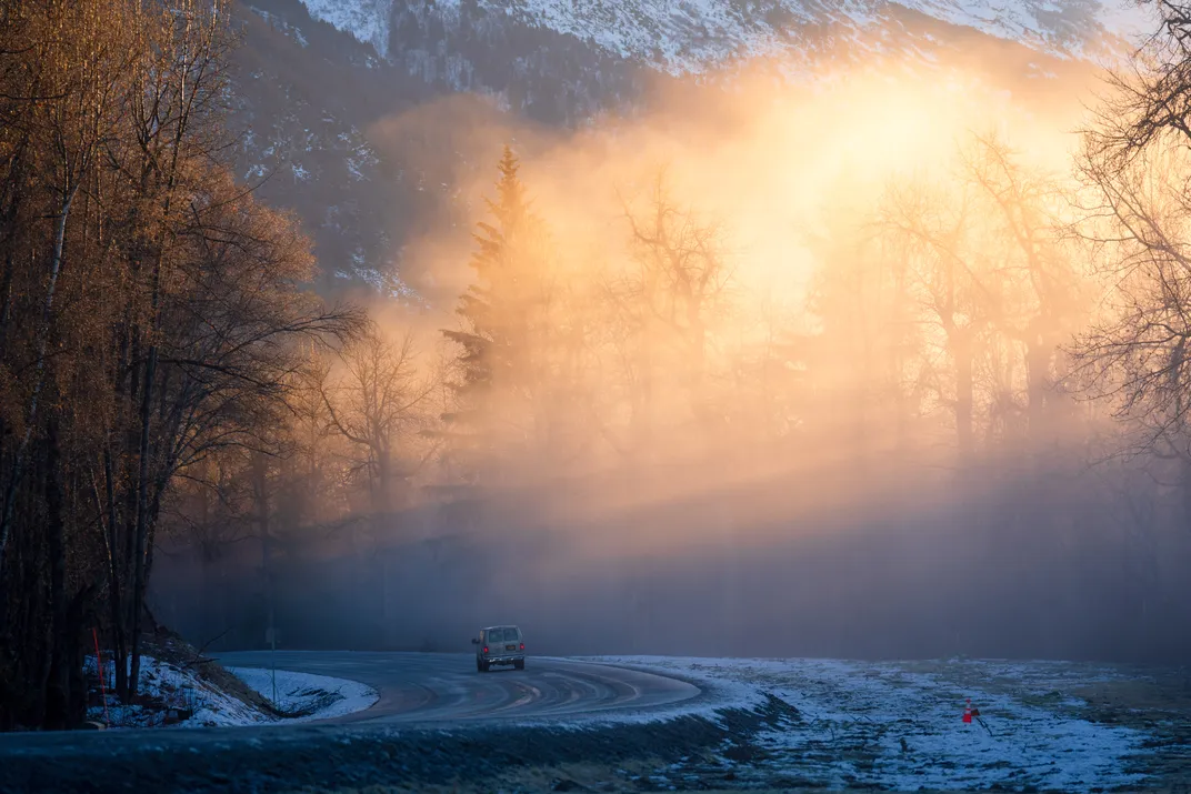 Snowcapped mountains can be seen beyond the fog and sunrays on a quiet road at dawn.
