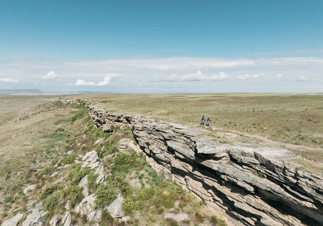 Two hikers walking beside a rugged cliff edge in an expansive prairie landscape.
