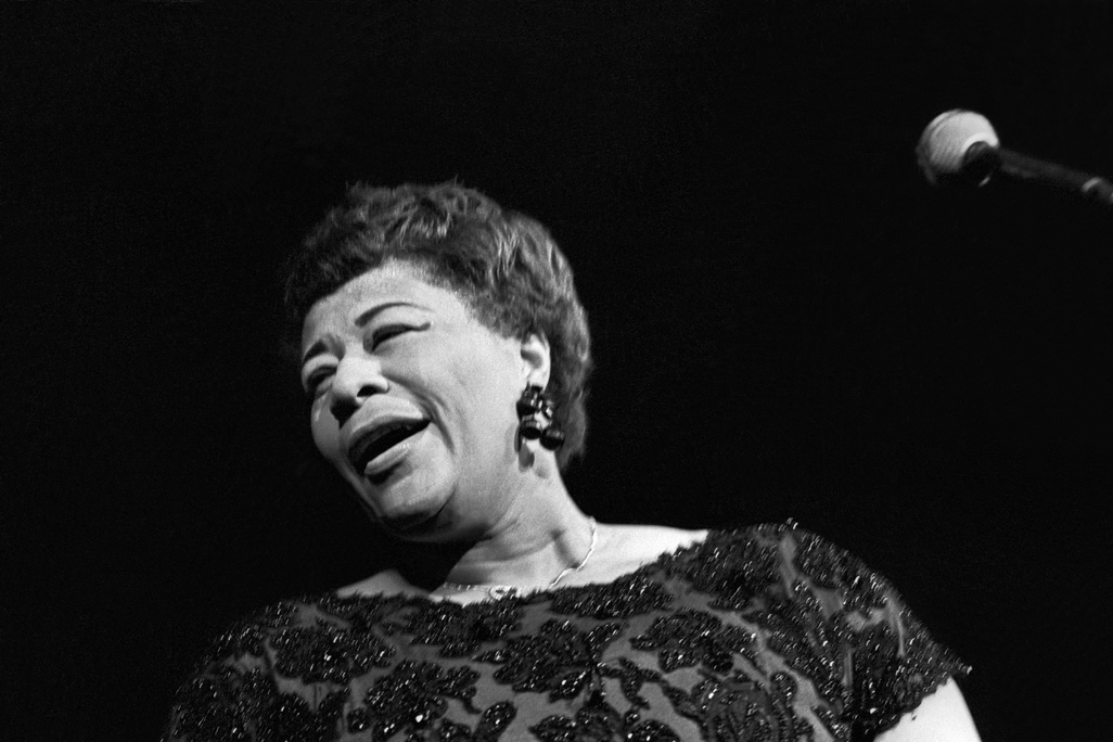 A black and white image, closely cropped to Fitzgerald's profile with the microphone just peeking in on the side; Fitzgerald is singing and turns away from the camera, smiling, mouth half-open and eyes closed, wearing a beaded dress and short cropped hair