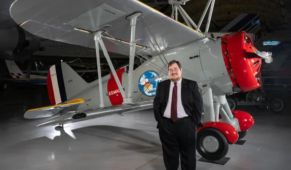 A white man in his 30s stands in front of a restored biplane displayed on a polished concrete floor inside a museum. He is wearing glasses and a dark suit with a white shirt and burgundy tie.