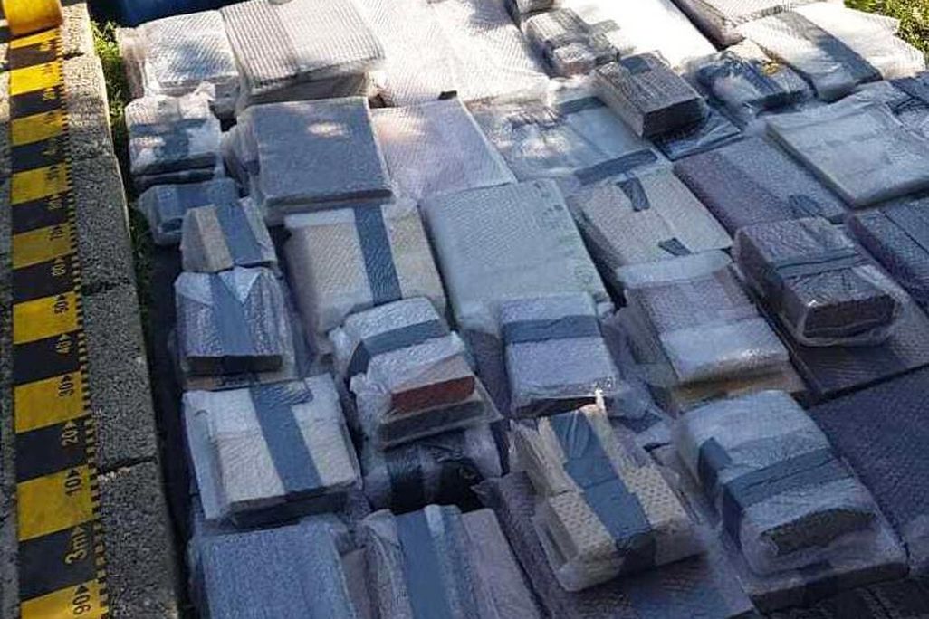 Stacks of books, which appear to be individually wrapped in protective fabric, lying on the ground in organized stacks next to a garbage bin and some plants