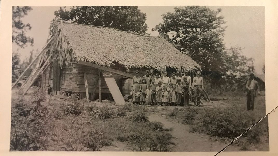 A group of people pose in front of a wooden building with thatched roof. Black-and-white photo with one corner ripped off.