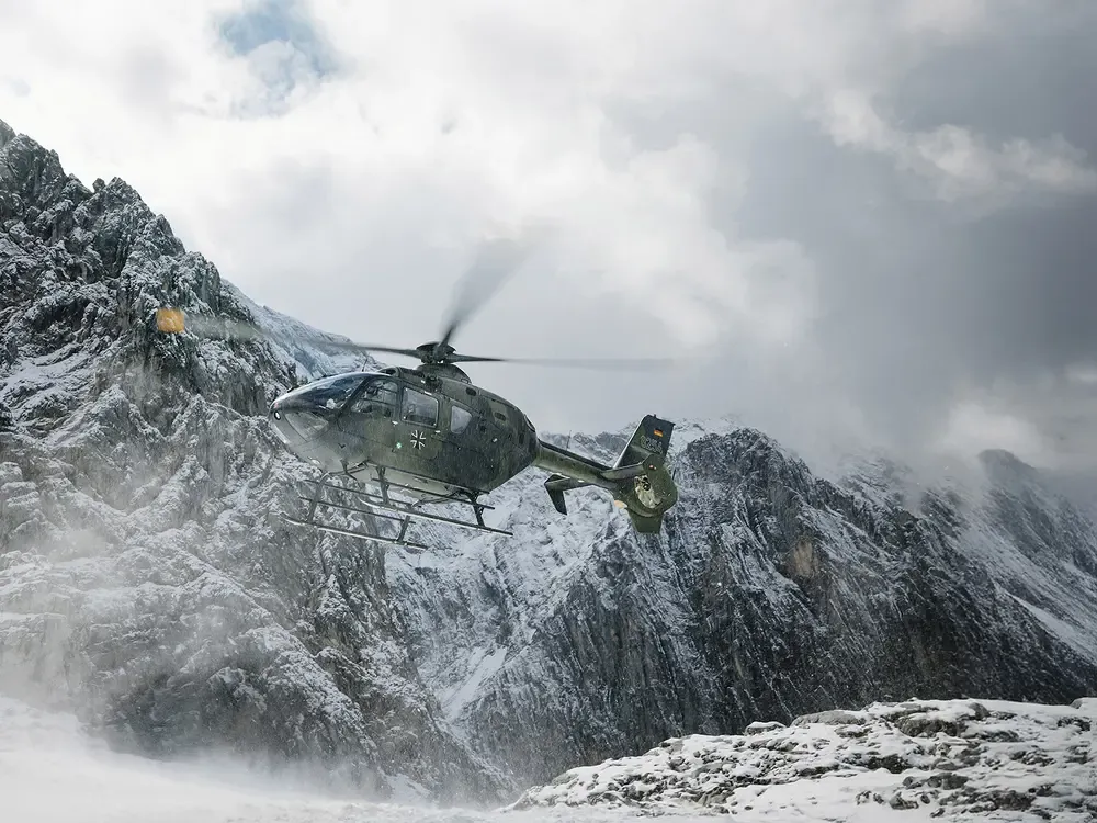 A helicopter flies low over a snow-covered, jagged mountain range. The rotors kick up a light mist of snow against the dark, rocky terrain.