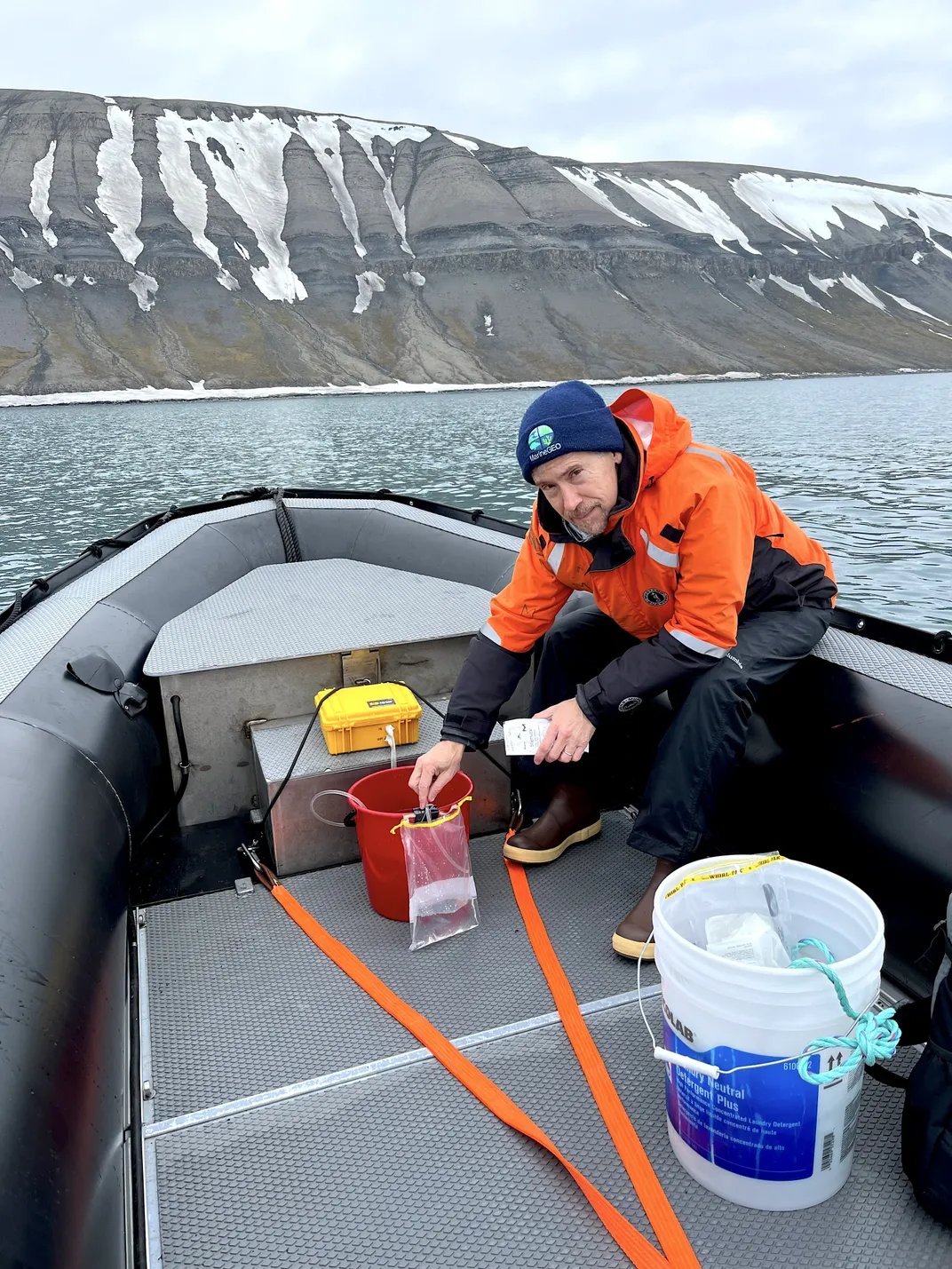 Man in orange jacket testing water sample in lifeboat, water around the boat, snowcapped mountains in the background