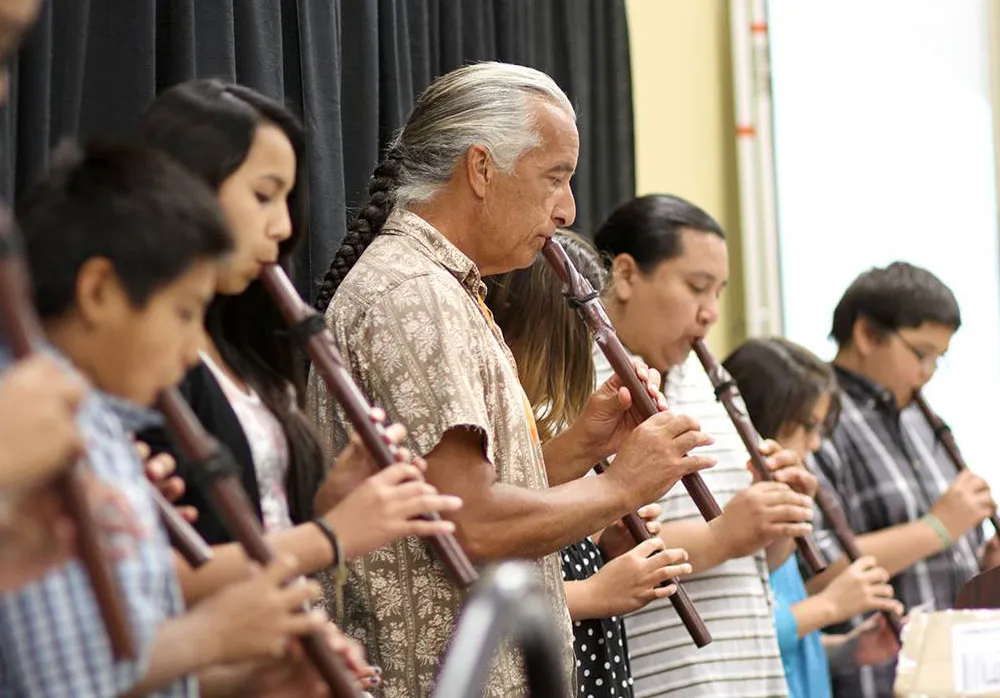 A row of people lines up playing flutes. Most are young students, and in the middle is an older man with a long gray braid.