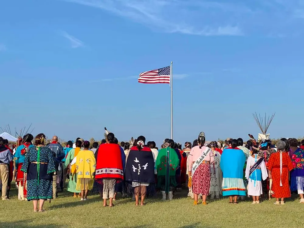 Members of the Kiowa Gourd Clan Ceremony stand as the flag of Spencer “Corky” Sahmaunt is raised. Carnegie, Oklahoma; July 4, 2019. Mr. Sahmaunt served in the U.S. Army during the Korean War and was a member of the Kiowa Black Leggings Warrior Society, as well as the Kiowa Gourd Clan.The Kiowa Flag Song, analogous to the Star Spangled Banner, accompanied the flag-raising. (Photo courtesy of Mari Frances Sahmaunt, used with permission)
