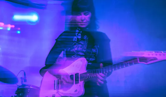 A woman with long dark hair and dark outfit plays a white electric guitar on a dimly blue lit stage. She and her guitar are slightly blurry.