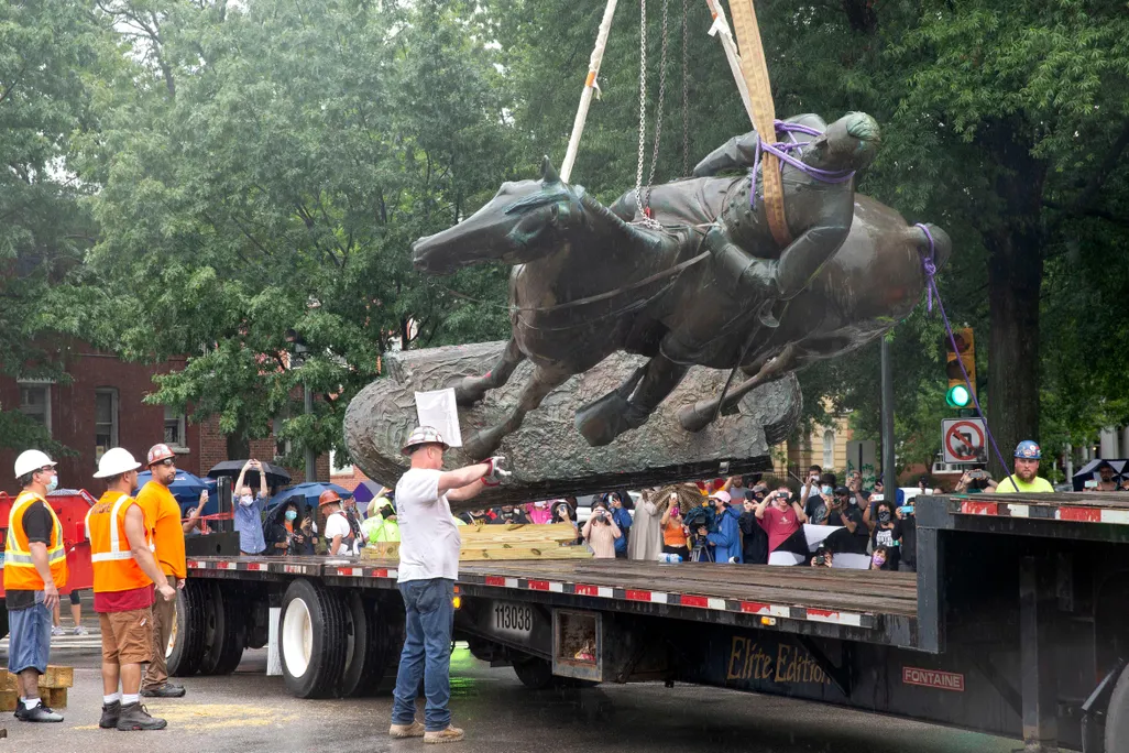 A group of people in hard hats, some in orange work vests, guide a large statue of a bearded man on a horse onto a truck's platform. The statue is dark gray larger-than-life, suspended by a rope and a crane (out of view); many onlookers watch nearby