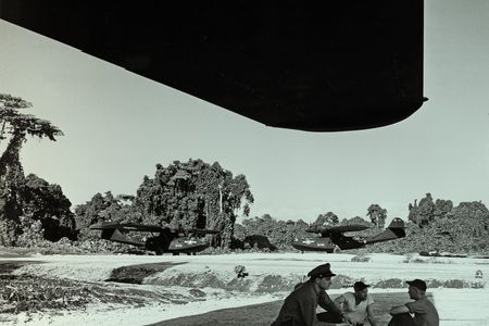 Three men sit under the shade of a massive airplane wing; one of the men is wearing an officer's hat. Two huge aircraft are parked in the background, both are painted black. Behind them is a lush island landscape. The photo has been given a green tint.