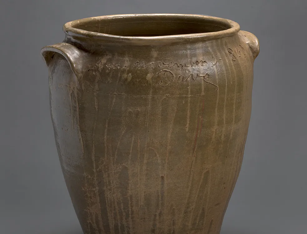 A brown, ceramic vessel with an inscription and the artist's name "Dave" carved around the opening.