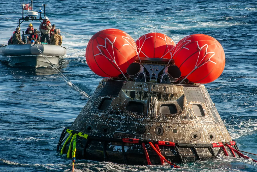 the top of the Orion capsule is seen above the water with some parachutes above it and recovery personnel in a boat behind it