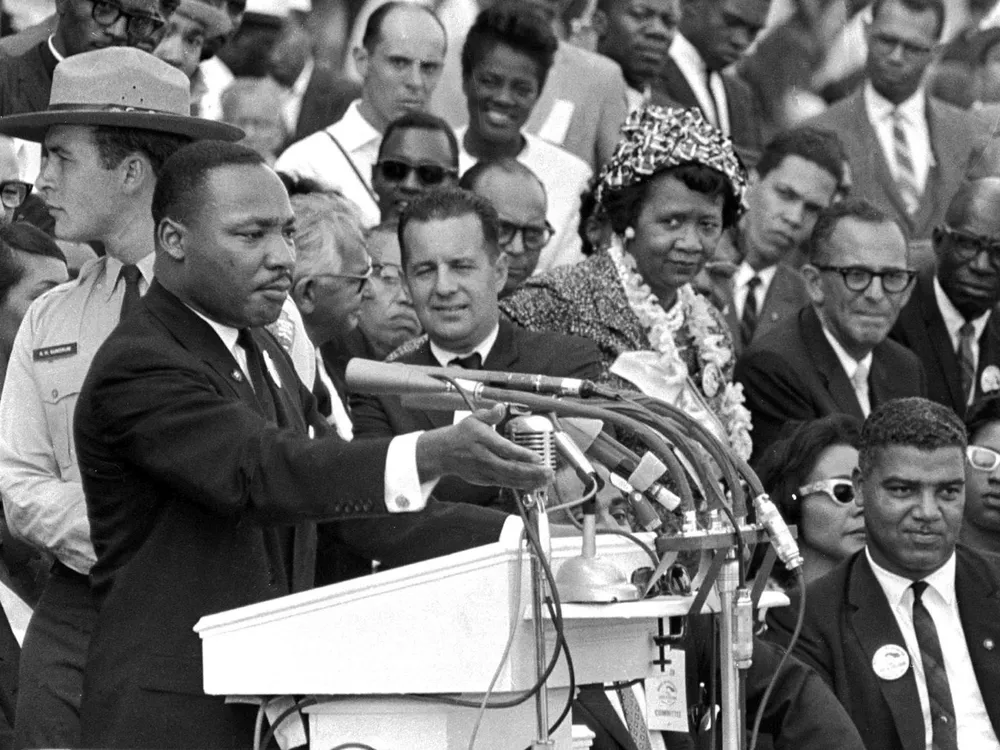 Martin Luther King, Jr. stands at a podium with several microphones. Behind him to his left is a crowd of seated and standing people, including Dorothy Height, turned towards him.