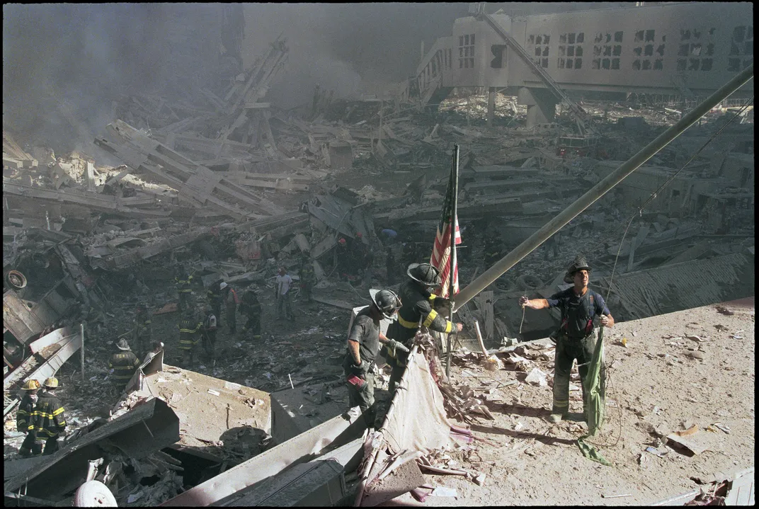 A group of three men stand in yellow evening light, casting shadows behind them, raising a flag in front of a sprawling scene of wreckage.