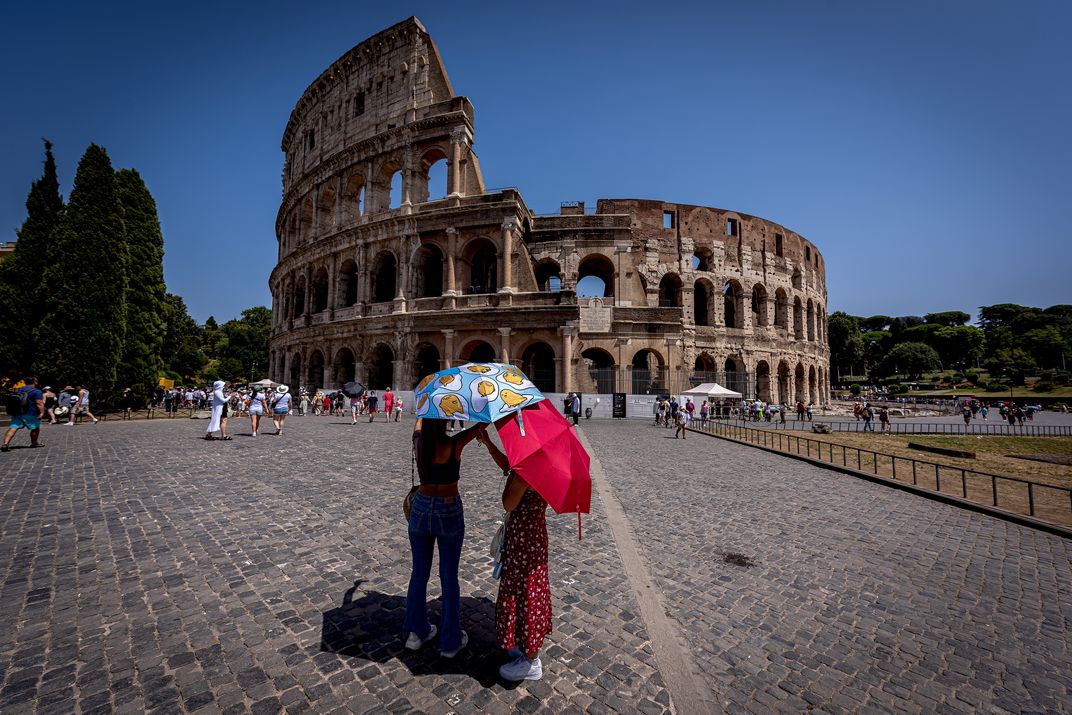 people with umbrellas stand in front of the colosseum on a cloudless day