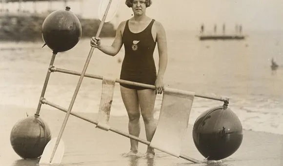 A black and white gelatin silver print of Gertrude Ederle standing on the shore with a 1920s flotation device.