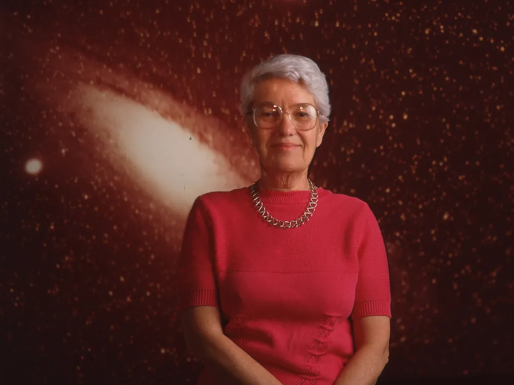 Vera Rubin stands against a backdrop of a star-filled galaxy with her hands loosely clasped in front of her.