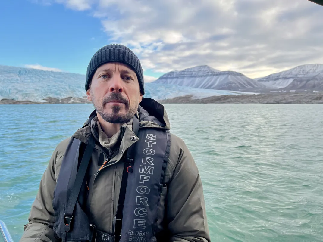 a man in a beanie sits with the ocean and glaciers in the background