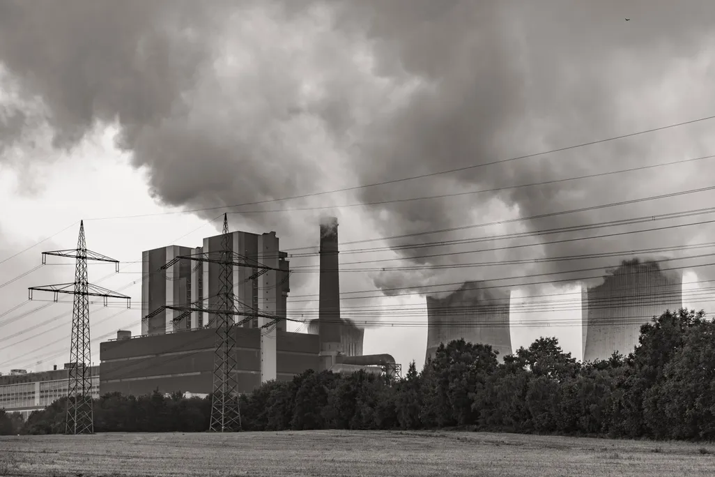 In black and white, power lines cross the foreground as a power plant billows smoke behind it.