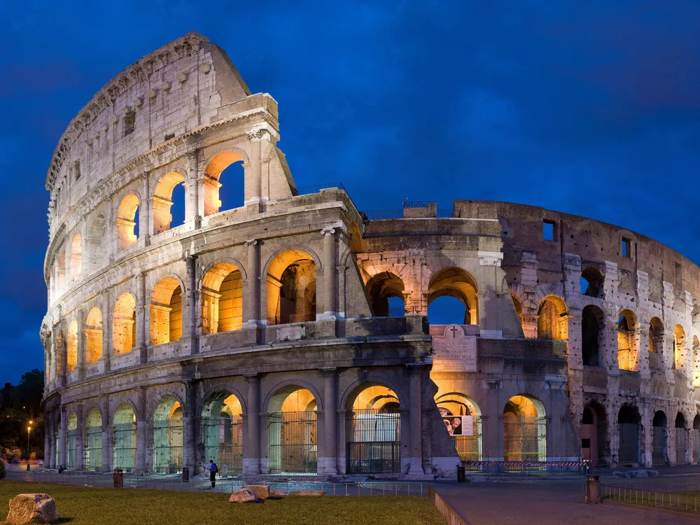 The Colosseum at night, with a blue sky and its arches lit up by yellow light. The structure is partially dismantled, with half its walls torn down