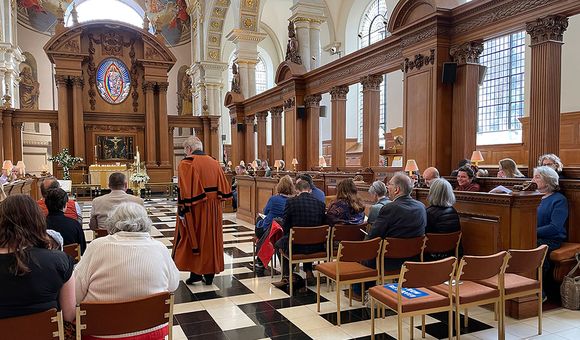 Interior of a church, with people seated in chairs and pews, and a person in reddish robe with black trim walking toward the pulpit.