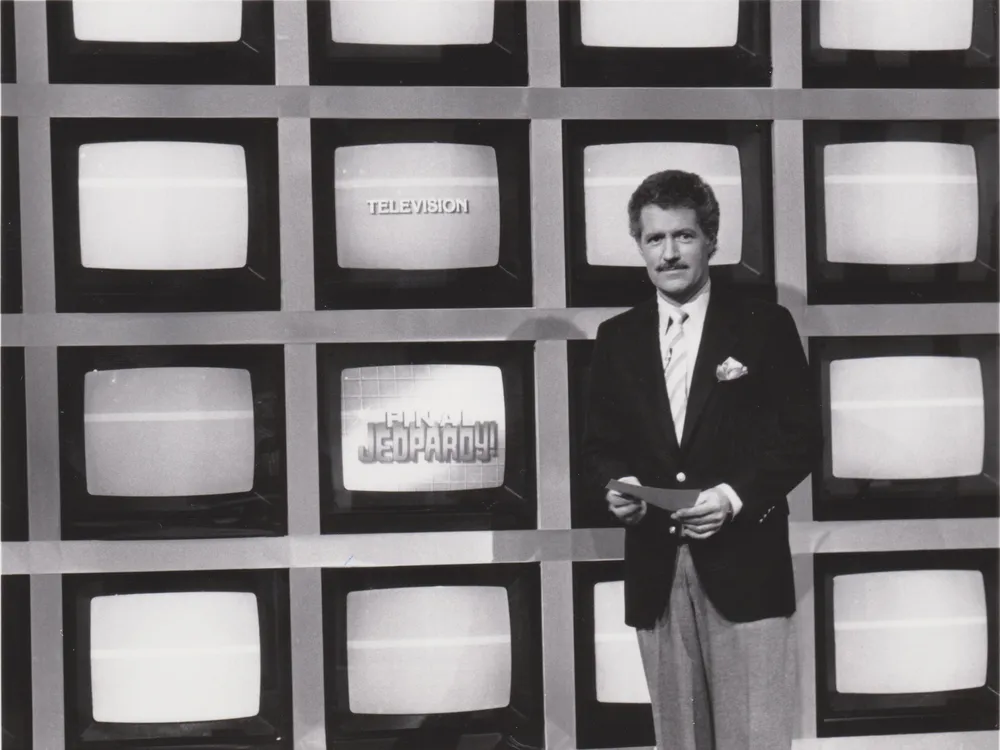 A black and white image of a young Trebek, a white man in a suit and tie with dark hair and a mustache, standing in front of a grid of TV screens